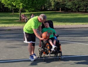 Photo of young man holding a basketball while sitting in a wheelchair. Coach is standing next to the young man and assisting.